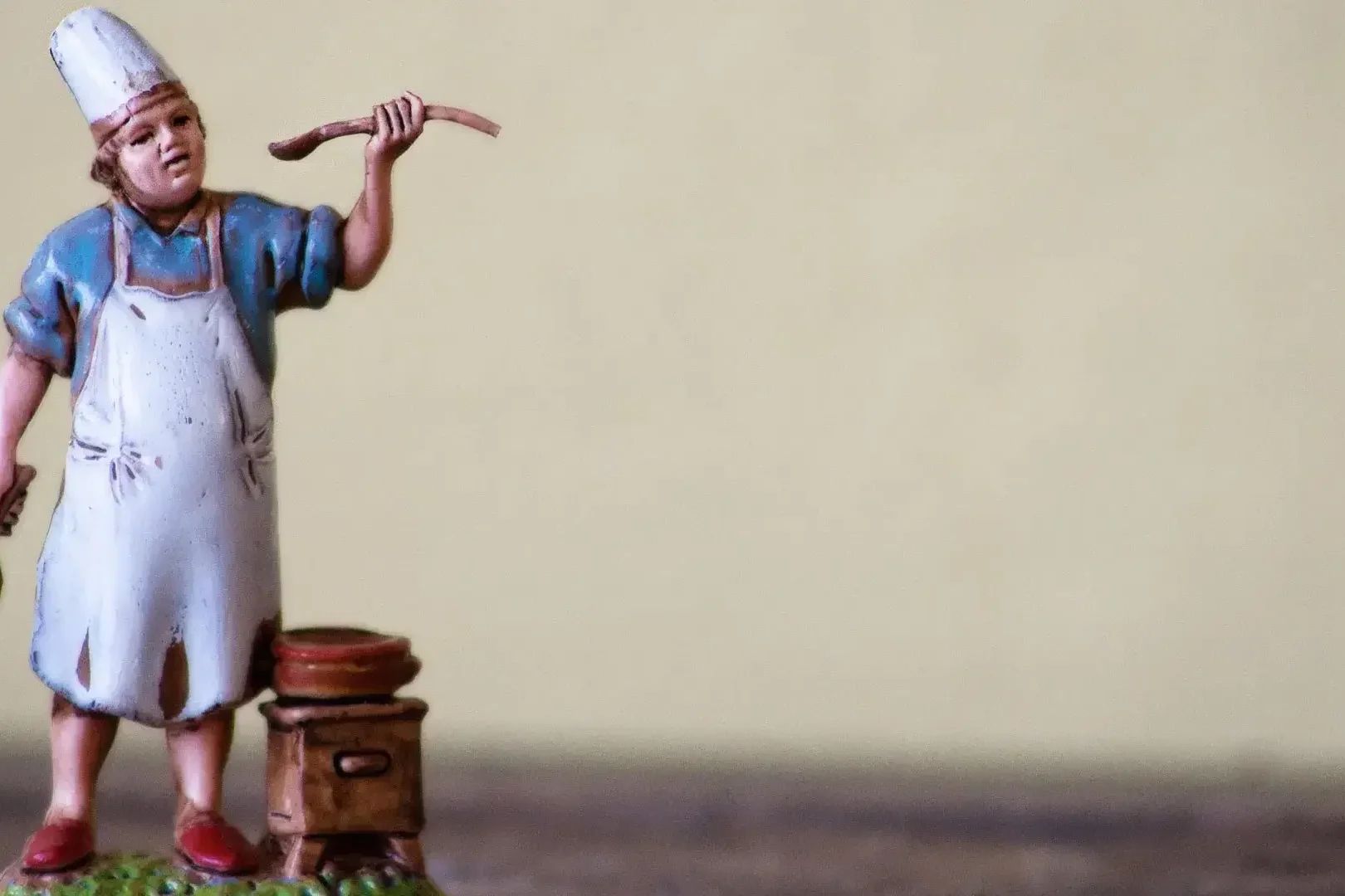 Una statuetta di un cuoco con cappello e grembiule bianco si trova accanto a una pentola su un fornello, con in mano un cucchiaio di legno e un mattarello: perfetto per chi sogna di assumere un cuoco personale Varese per creare un menù su misura.