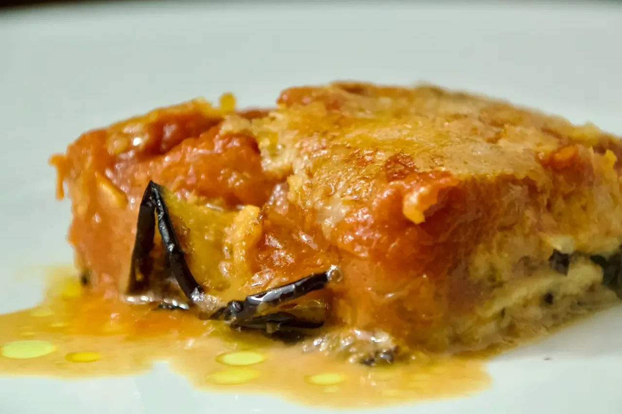 Primo piano di una fetta di parmigiana di melanzane con strati di salsa di pomodoro, formaggio fuso e melanzane al forno su un piatto bianco, presentata da Giorgio di Cucino di Te, chef a domicilio a Varese.