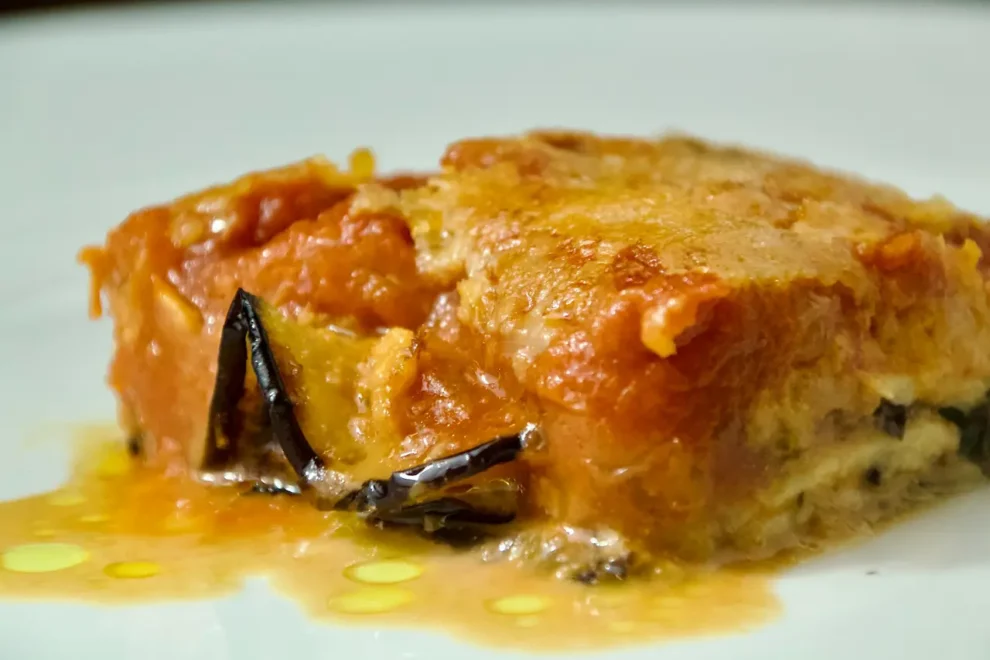 Primo piano di una fetta di parmigiana di melanzane con strati di salsa di pomodoro, formaggio fuso e melanzane al forno su un piatto bianco, presentata da Giorgio di Cucino di Te, chef a domicilio a Varese.